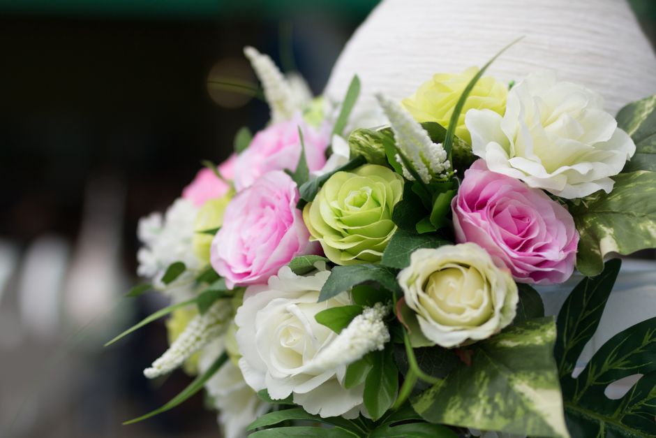 A floral arrangement featuring a bouquet of fresh roses and lisianthus flowers in soft pink, white, and pale green, with lush green foliage and small white accent flowers, set against a blurred backgr