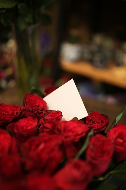 A close-up view of a bouquet of fresh red roses with tightly layered petals, arranged with green stems and leaves, resting on a surface inside a flower shop. A plain white card is tucked among the ros