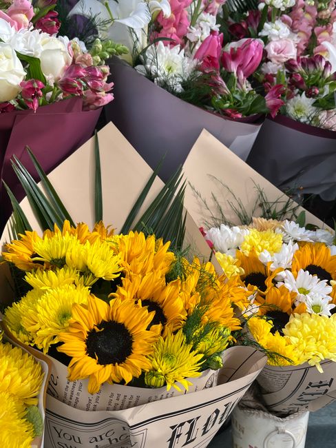 A vibrant bouquet of fresh sunflowers with large, bright yellow petals and dark centers, accompanied by green foliage and long, pointed leaves, wrapped in newspaper-style paper. The arrangement is pos