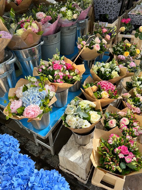 A display of fresh floral arrangements and bouquets featuring pink, white, and blue flowers, including roses, hydrangeas, and carnations, wrapped in brown paper or placed in metallic and ceramic pots.