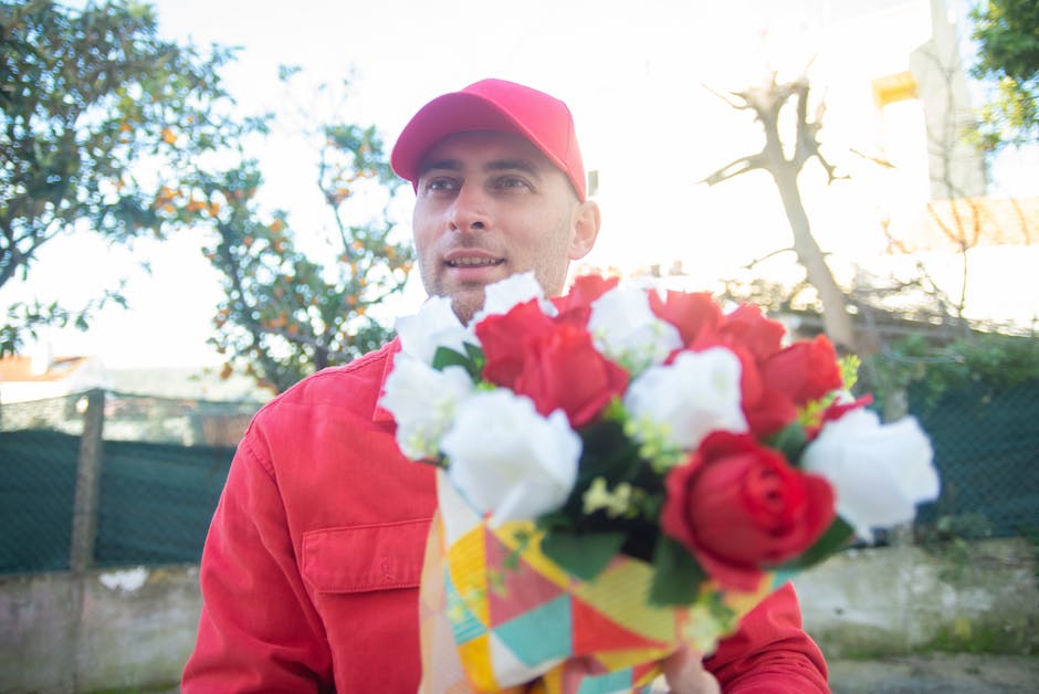 A male flower delivery courier, dressed in a red uniform and matching cap, is outdoors holding a vibrant bouquet of fresh flowers, including red and white blossoms, wrapped in decorative patterned pap
